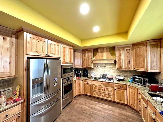 kitchen with wood-type flooring, a raised ceiling, stainless steel appliances, custom range hood, and decorative backsplash