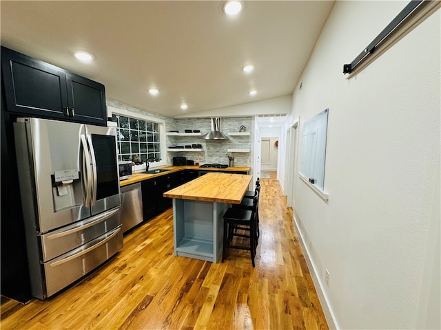 kitchen featuring wall chimney range hood, appliances with stainless steel finishes, a breakfast bar, wood counters, and a center island