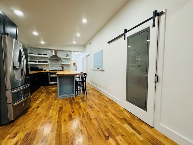 kitchen with a breakfast bar area, appliances with stainless steel finishes, a barn door, butcher block counters, and wall chimney exhaust hood