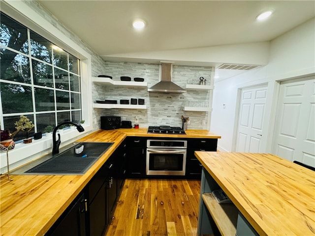 kitchen with decorative backsplash, butcher block counters, stainless steel oven, wall chimney exhaust hood, and sink