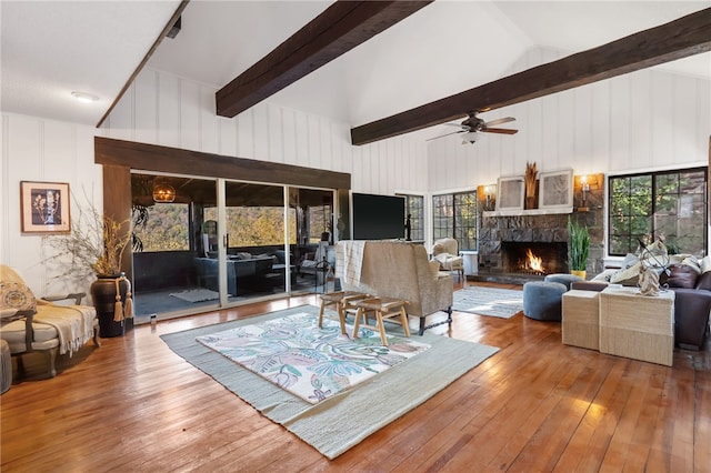 living room with ceiling fan, wood-type flooring, high vaulted ceiling, beamed ceiling, and a stone fireplace