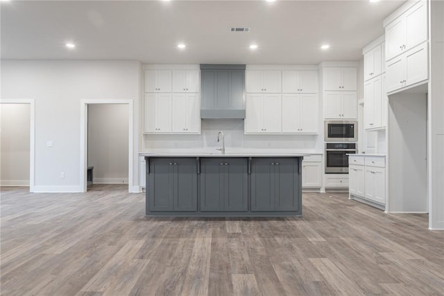 kitchen with built in microwave, stainless steel oven, gray cabinets, a kitchen island with sink, and white cabinets