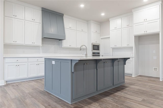 kitchen with light hardwood / wood-style floors, white cabinetry, built in microwave, and an island with sink