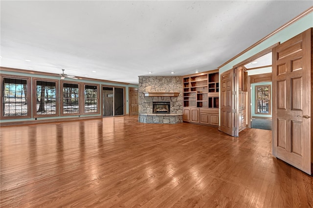 unfurnished living room featuring wood-type flooring, a stone fireplace, ceiling fan, ornamental molding, and built in shelves