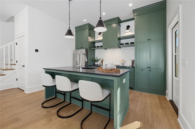 kitchen featuring green cabinetry, decorative backsplash, hanging light fixtures, a kitchen island with sink, and light hardwood / wood-style flooring