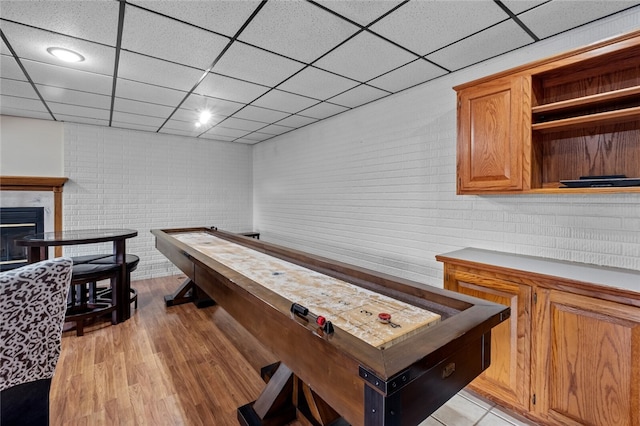 rec room with light wood finished floors, brick wall, a drop ceiling, and a fireplace