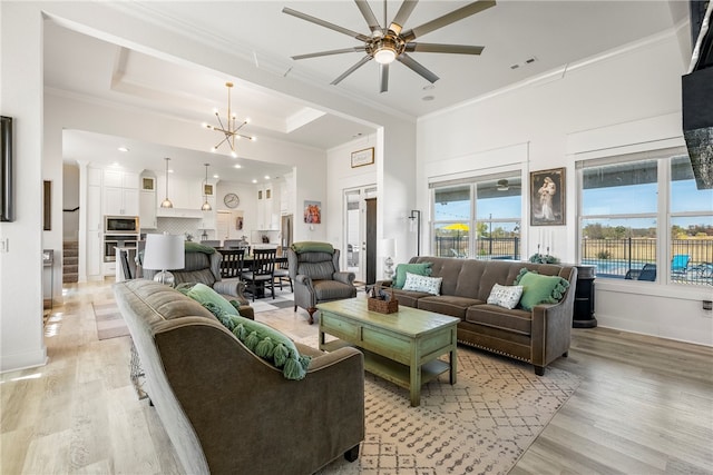 living room with crown molding, ceiling fan with notable chandelier, light wood-type flooring, and a raised ceiling