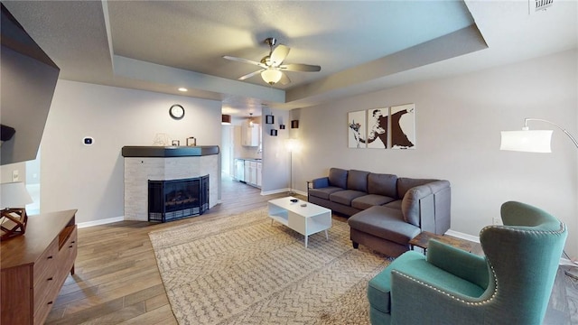 living room featuring a raised ceiling, wood-type flooring, a tile fireplace, and ceiling fan