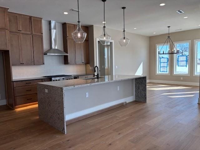 kitchen with a kitchen island with sink, a sink, light stone countertops, wall chimney exhaust hood, and decorative light fixtures