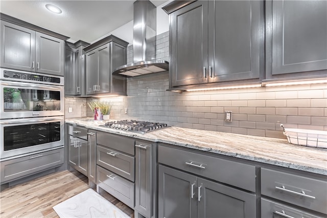 kitchen featuring light stone countertops, wall chimney exhaust hood, tasteful backsplash, appliances with stainless steel finishes, and light wood-type flooring