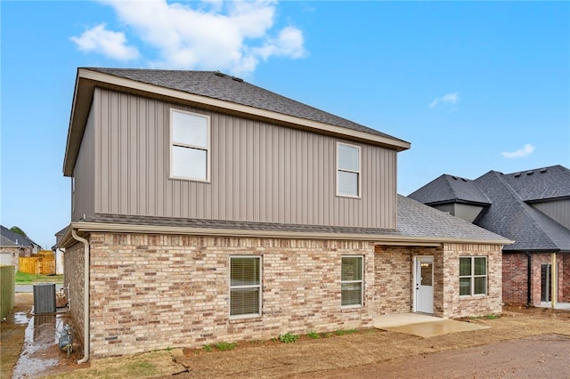 back of property with a shingled roof, cooling unit, board and batten siding, and brick siding