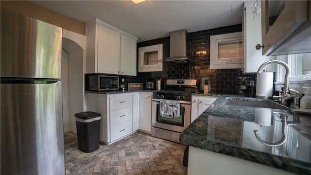 kitchen with backsplash, stainless steel appliances, wall chimney range hood, dark stone countertops, and white cabinetry