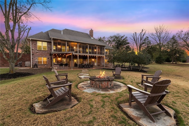 back house at dusk featuring a sunroom, a patio area, an outdoor fire pit, and a lawn