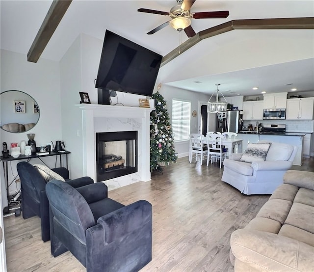 living room featuring sink, vaulted ceiling with beams, a high end fireplace, light hardwood / wood-style floors, and ceiling fan with notable chandelier