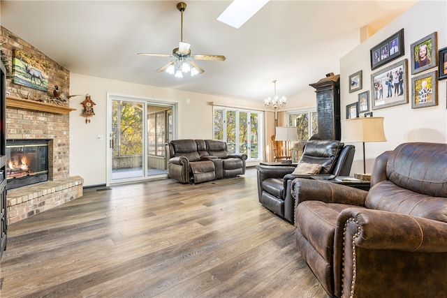 living room with a brick fireplace, ceiling fan with notable chandelier, hardwood / wood-style flooring, and lofted ceiling with skylight