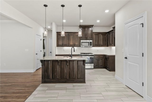 kitchen featuring appliances with stainless steel finishes, a kitchen island with sink, hanging light fixtures, dark brown cabinetry, and light stone countertops