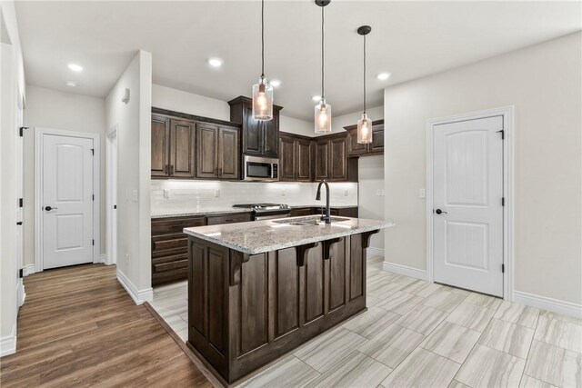 kitchen featuring dark brown cabinetry, sink, light stone counters, appliances with stainless steel finishes, and an island with sink