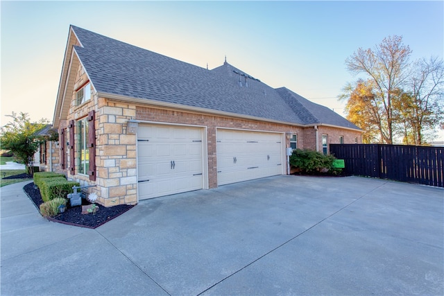 property exterior at dusk featuring an attached garage, a shingled roof, fence, and concrete driveway