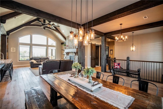 dining space featuring vaulted ceiling with beams, wood-type flooring, a large fireplace, ceiling fan with notable chandelier, and a barn door