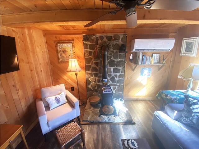 living area featuring ceiling fan, beam ceiling, an AC wall unit, hardwood / wood-style floors, and a wood stove