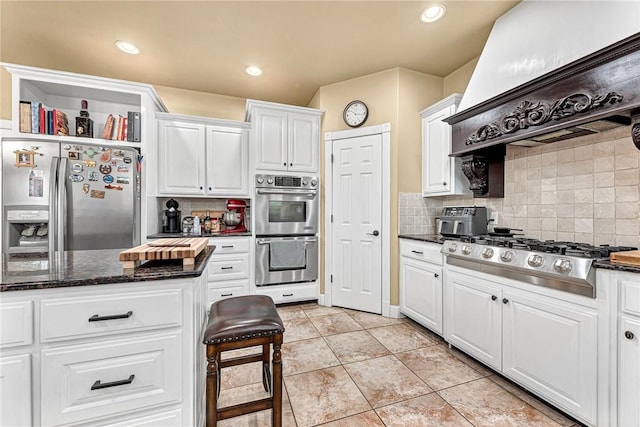 kitchen featuring backsplash, premium range hood, stainless steel appliances, dark stone countertops, and white cabinets