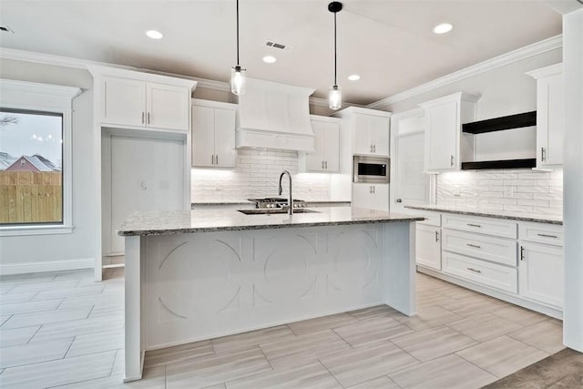 kitchen featuring light stone countertops, premium range hood, pendant lighting, a center island with sink, and stainless steel microwave