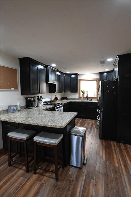 kitchen featuring stainless steel gas range oven, dark wood-type flooring, black refrigerator, light stone countertops, and kitchen peninsula