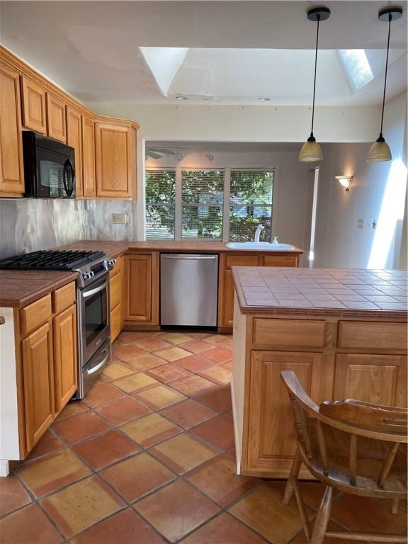 kitchen with tile countertops, a skylight, hanging light fixtures, appliances with stainless steel finishes, and backsplash