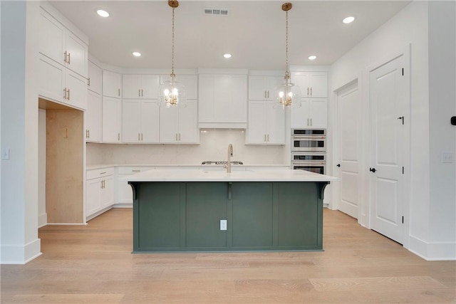 kitchen featuring double oven, an island with sink, light wood-type flooring, light countertops, and white cabinets