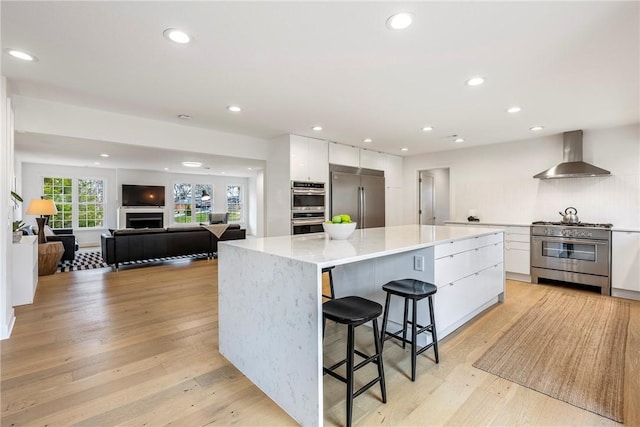 kitchen with wall chimney range hood, light hardwood / wood-style flooring, a spacious island, white cabinets, and high end appliances