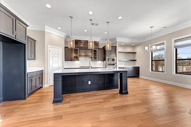 kitchen with an inviting chandelier, ornamental molding, an island with sink, light wood-type flooring, and appliances with stainless steel finishes