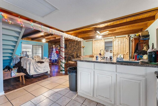 kitchen with ceiling fan, light tile patterned floors, beam ceiling, white cabinets, and a stone fireplace