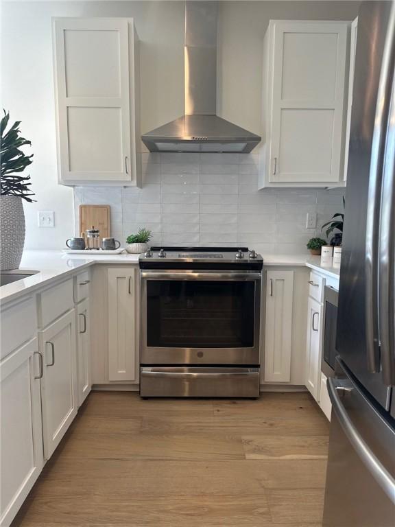 kitchen with white cabinetry, wall chimney range hood, decorative backsplash, appliances with stainless steel finishes, and light wood-type flooring