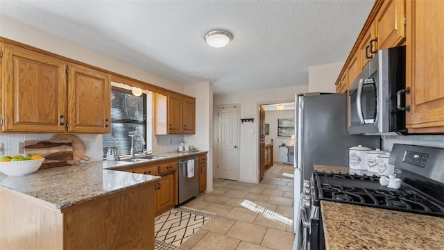 kitchen featuring a textured ceiling, a sink, appliances with stainless steel finishes, brown cabinets, and light stone countertops