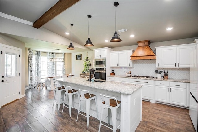 kitchen with custom exhaust hood, stainless steel appliances, vaulted ceiling with beams, white cabinetry, and an island with sink