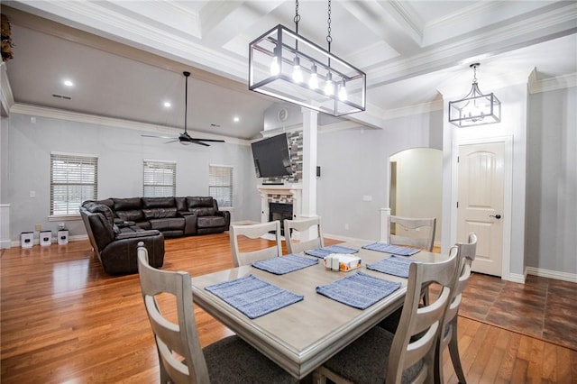 dining room featuring ceiling fan with notable chandelier, hardwood / wood-style flooring, a stone fireplace, and crown molding