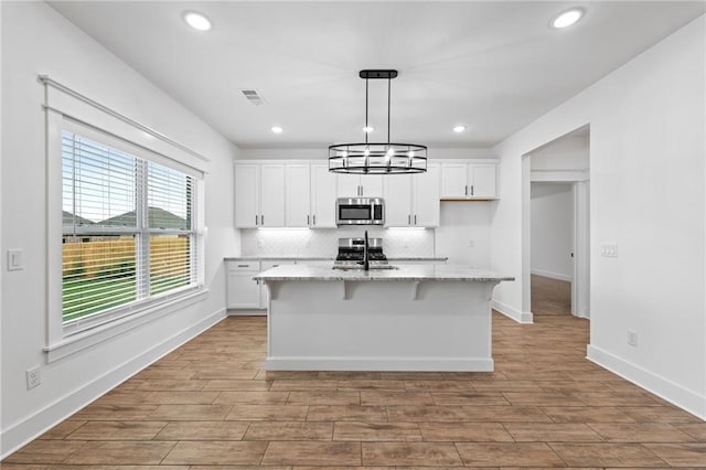 kitchen with light stone countertops, stainless steel appliances, decorative light fixtures, a center island with sink, and white cabinets