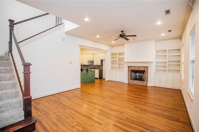 unfurnished living room with a brick fireplace, ornamental molding, ceiling fan, hardwood / wood-style flooring, and built in features