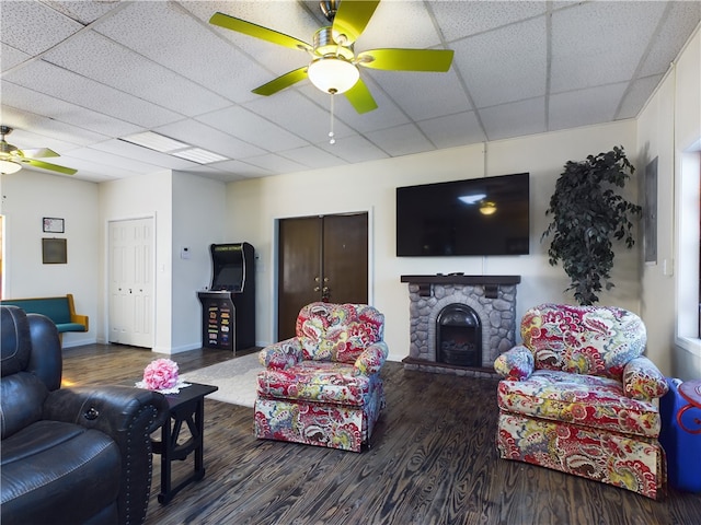 living room with a drop ceiling, ceiling fan, a stone fireplace, and dark wood-type flooring