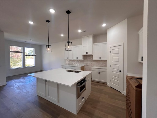 kitchen featuring dark hardwood / wood-style flooring, stainless steel oven, a center island with sink, white cabinets, and hanging light fixtures