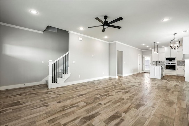 unfurnished living room with ceiling fan with notable chandelier, sink, light wood-type flooring, and crown molding