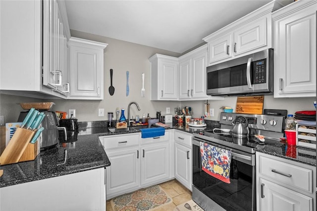 kitchen with appliances with stainless steel finishes, sink, light tile patterned floors, dark stone countertops, and white cabinetry