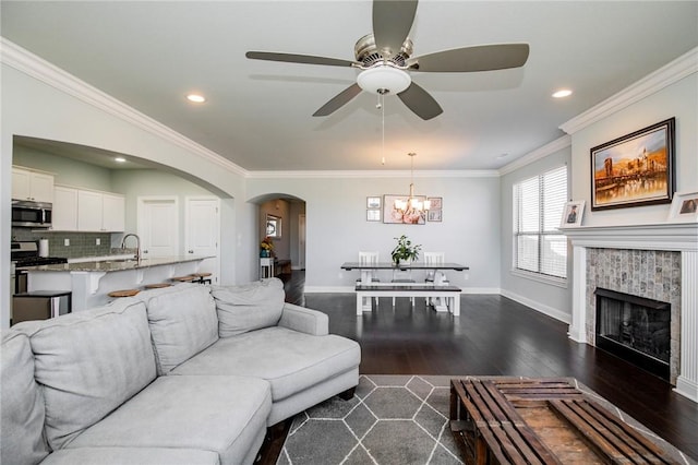 living room featuring dark hardwood / wood-style flooring, ceiling fan with notable chandelier, a tiled fireplace, ornamental molding, and sink