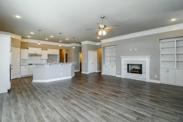 unfurnished living room with a tiled fireplace, ceiling fan, built in shelves, and crown molding