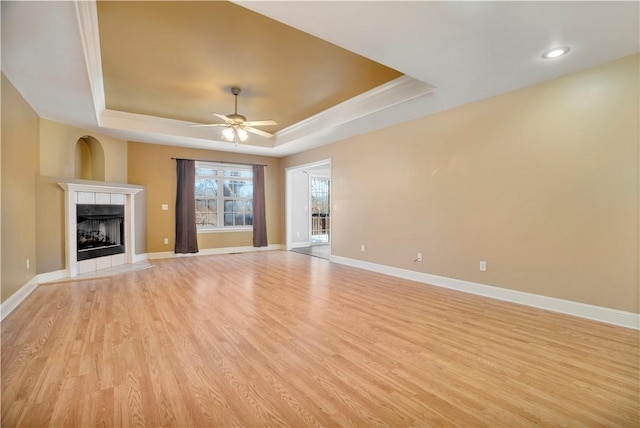 unfurnished living room with a tiled fireplace, a tray ceiling, light wood-style floors, and baseboards