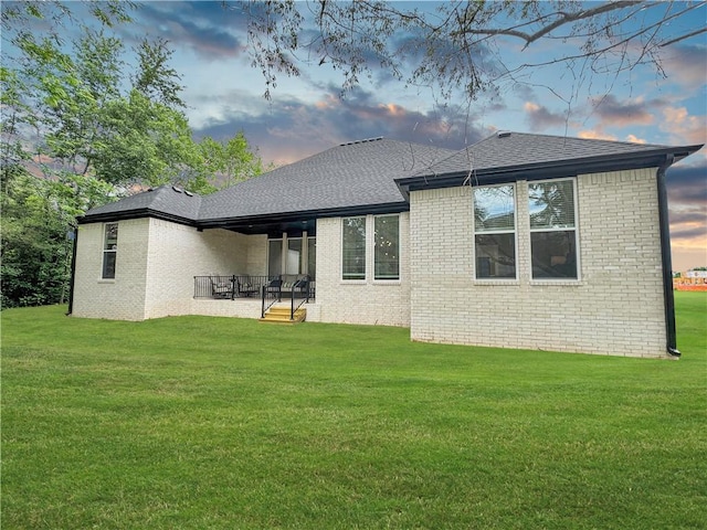 back of house at dusk featuring a yard, brick siding, a patio area, and a shingled roof