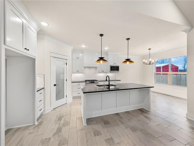 kitchen with hanging light fixtures, stainless steel microwave, sink, white cabinetry, and a kitchen island with sink