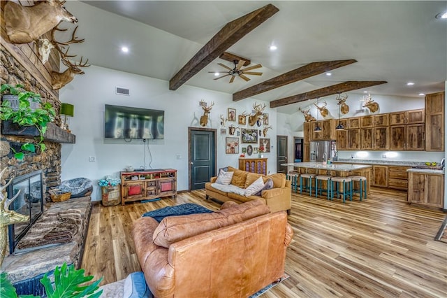 living room featuring lofted ceiling with beams, a stone fireplace, visible vents, a ceiling fan, and light wood finished floors