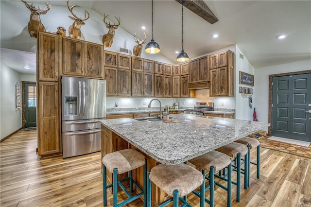 kitchen with light stone counters, a sink, hanging light fixtures, appliances with stainless steel finishes, and an island with sink