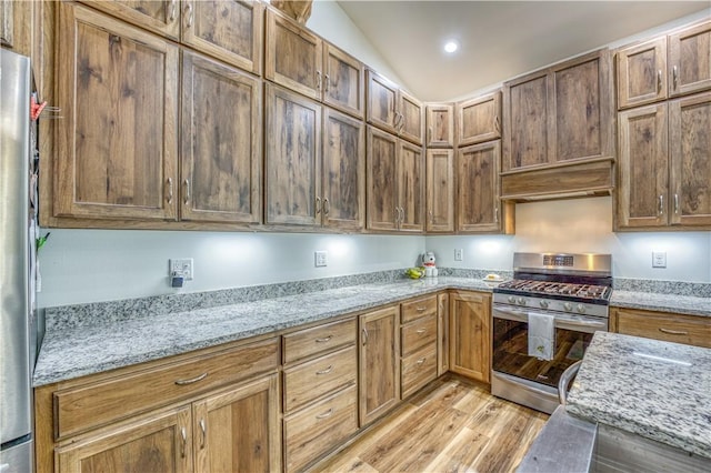 kitchen with brown cabinetry, light stone counters, range hood, vaulted ceiling, and stainless steel appliances